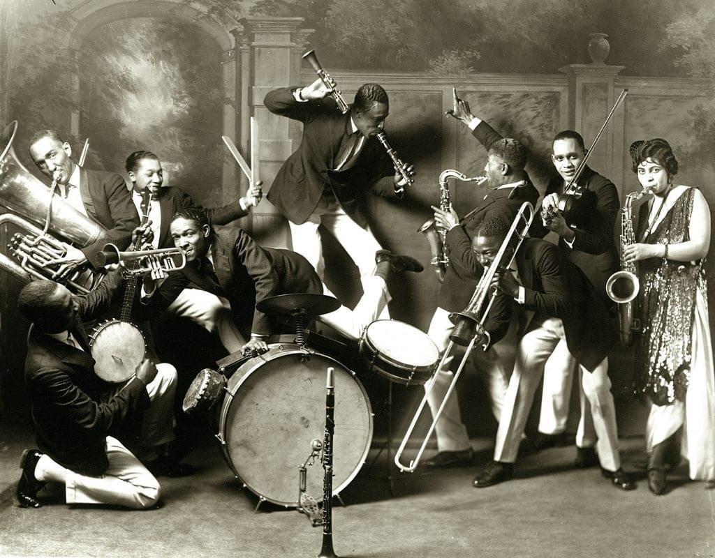A lively jazz band with nine musicians play various instruments, including trumpet, saxophone, drums, clarinet, and tuba, in an energetic pose against an ornate painted backdrop. One woman stands at the far right.