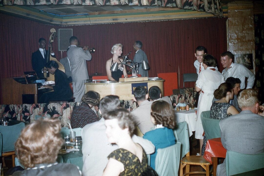 A lively nightclub scene with a jazz band performing on stage, including a female singer at the microphone. Well-dressed guests are seated at tables, talking and enjoying the music in a warmly lit, retro-style venue.