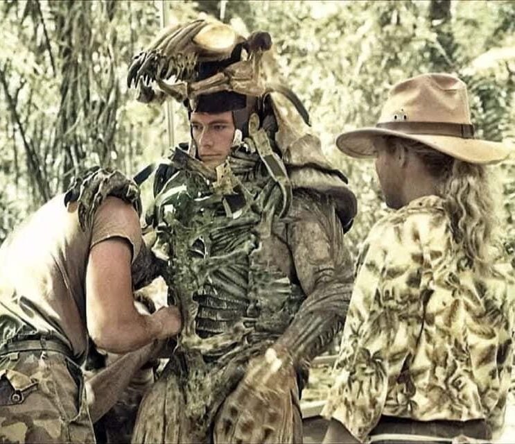 Three people in a jungle setting: one is wearing an alien costume, another is adjusting it, and a third person in a hat watches. The letters "JCVD" appear in the top left corner.