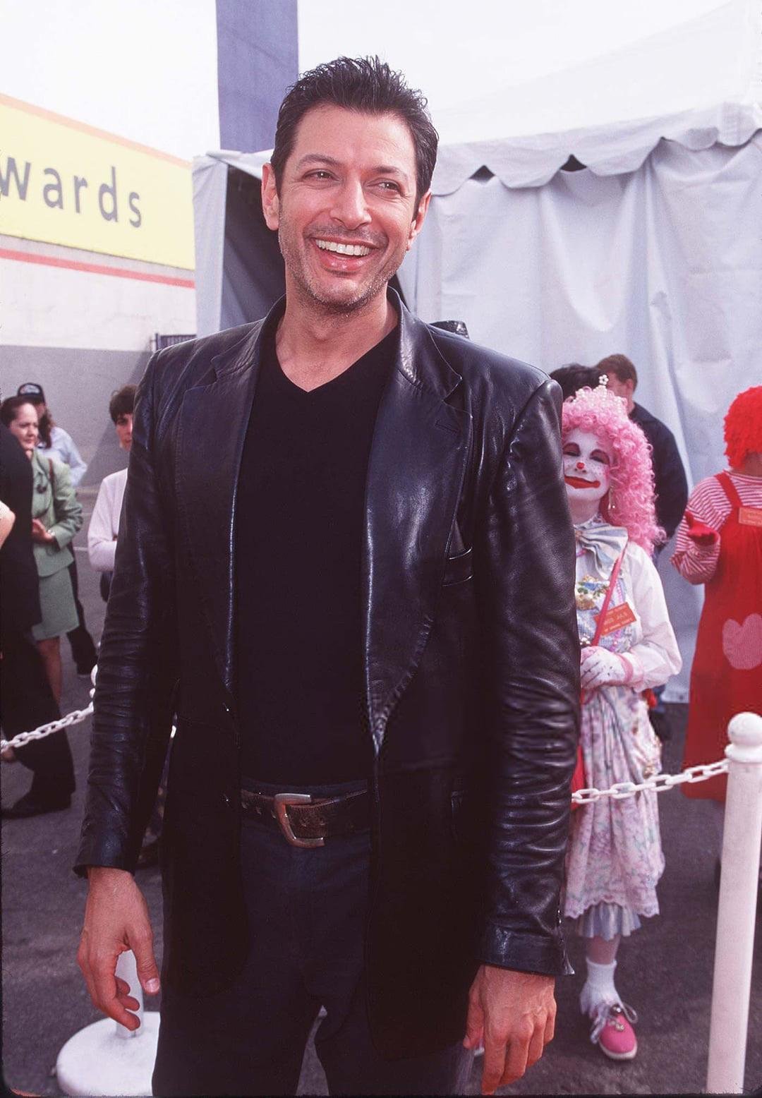 A smiling man in a black leather jacket and black shirt stands outdoors near a red rope barrier, with people and two clowns, one in a pink wig and white face paint, in the background.