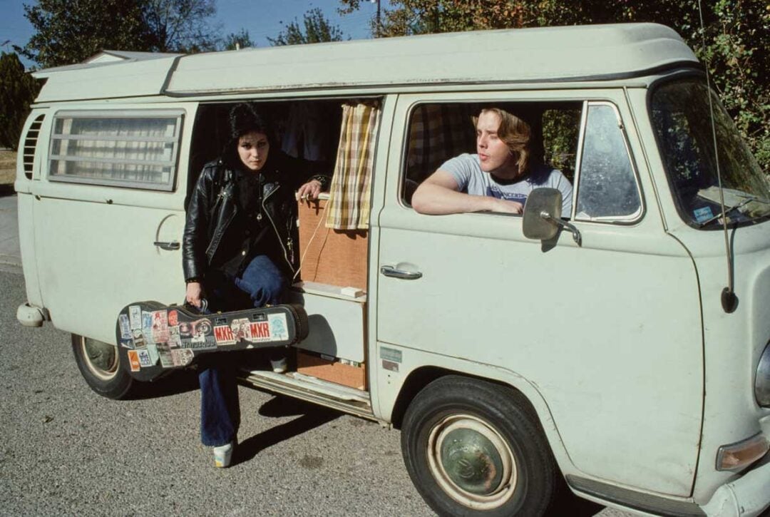 A person in a black leather jacket holds a guitar case and stands beside an open van door, while another person leans out from inside the vintage white van, parked on a sunny street.