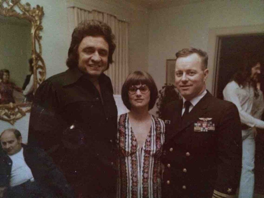 Three people pose indoors: a man in a black shirt, a woman with glasses and a patterned dress, and a man in a military uniform. Other people are visible in the background, including a seated man and a person in white.