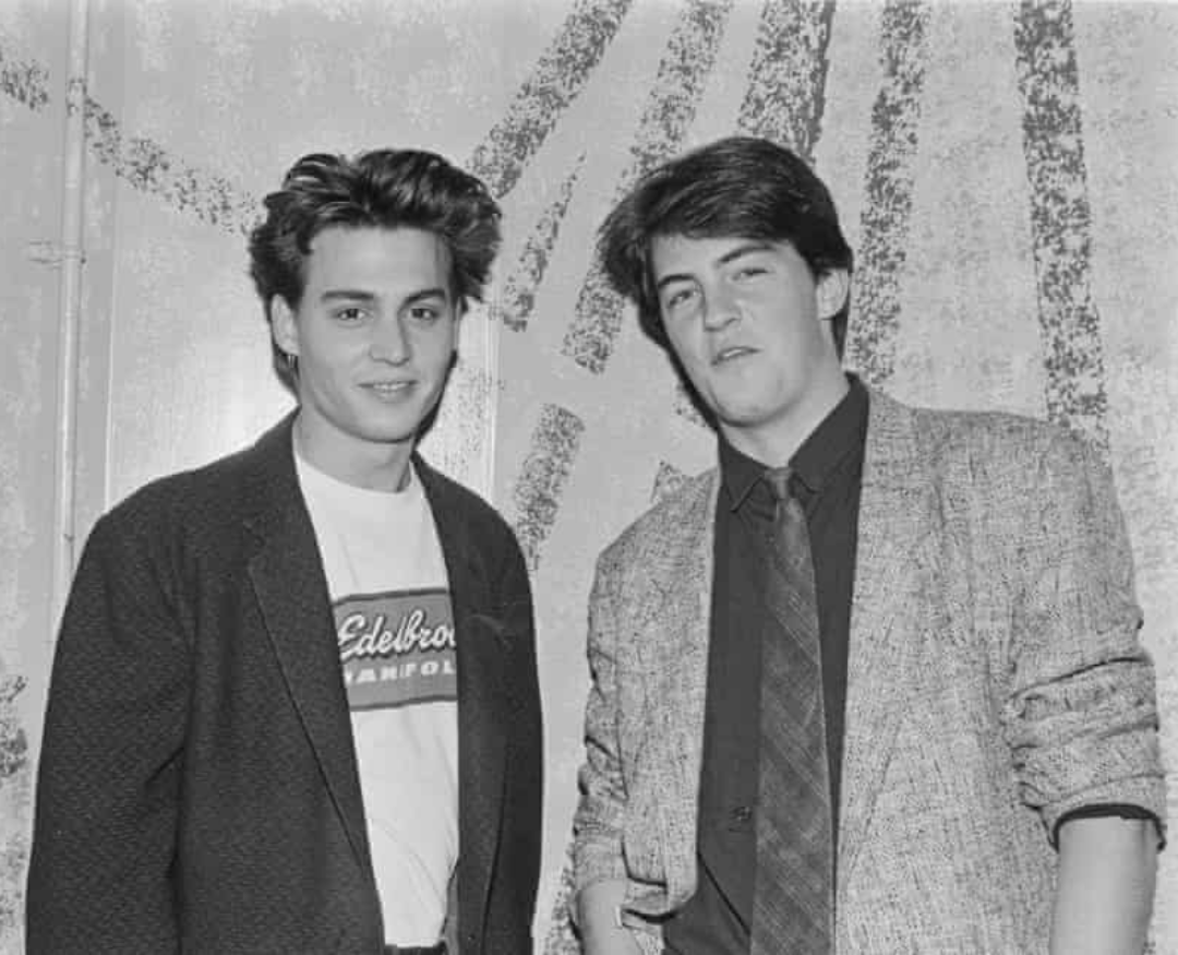 Two young men stand side by side, both wearing jackets. The man on the left has tousled hair and a white graphic T-shirt; the man on the right wears a tie and patterned blazer. They are smiling slightly in front of an abstract backdrop.