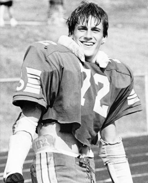 A young football player wearing a cropped jersey with the number 72 smiles on a field. He has wet hair, padded shoulders, and arm wraps, with a blurred background of grass and a track. The photo is in black and white.