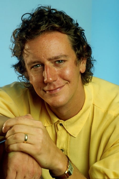 A man with curly brown hair wearing a yellow shirt smiles gently at the camera. He is resting his arms on a surface, showing a gold ring on his finger and a watch on his wrist. The background is light blue.