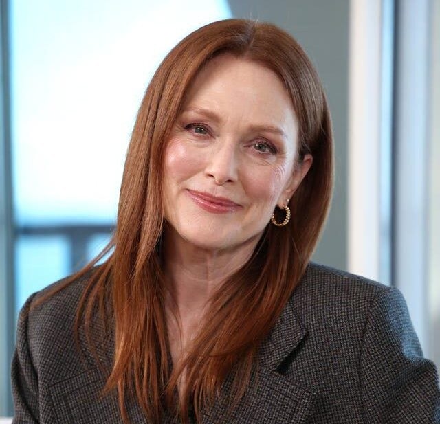 A woman with long, straight red hair and fair skin smiles softly. She is wearing a dark textured blazer and gold hoop earrings, seated indoors with a blurred background.