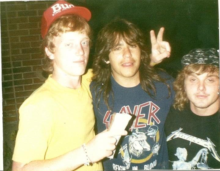 Three young men pose together; one wears a red cap and yellow shirt, another a "Slayer" shirt and gives a peace sign, and the third wears a cap and "Metallica" shirt. They stand in front of a brick wall at night.