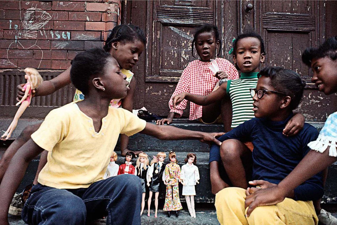 Six children sit and play with Barbie dolls on a set of worn outdoor steps in front of a brick building. The children appear engaged and lively, surrounded by several dolls lined up on a step.