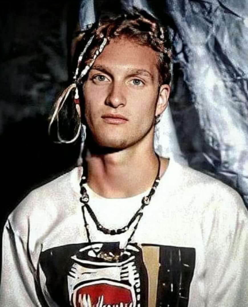 A young man with light skin and braided blond hair, some braids decorated with beads, wears a white graphic t-shirt and layered necklaces. He stands in front of a draped fabric background, looking directly at the camera.