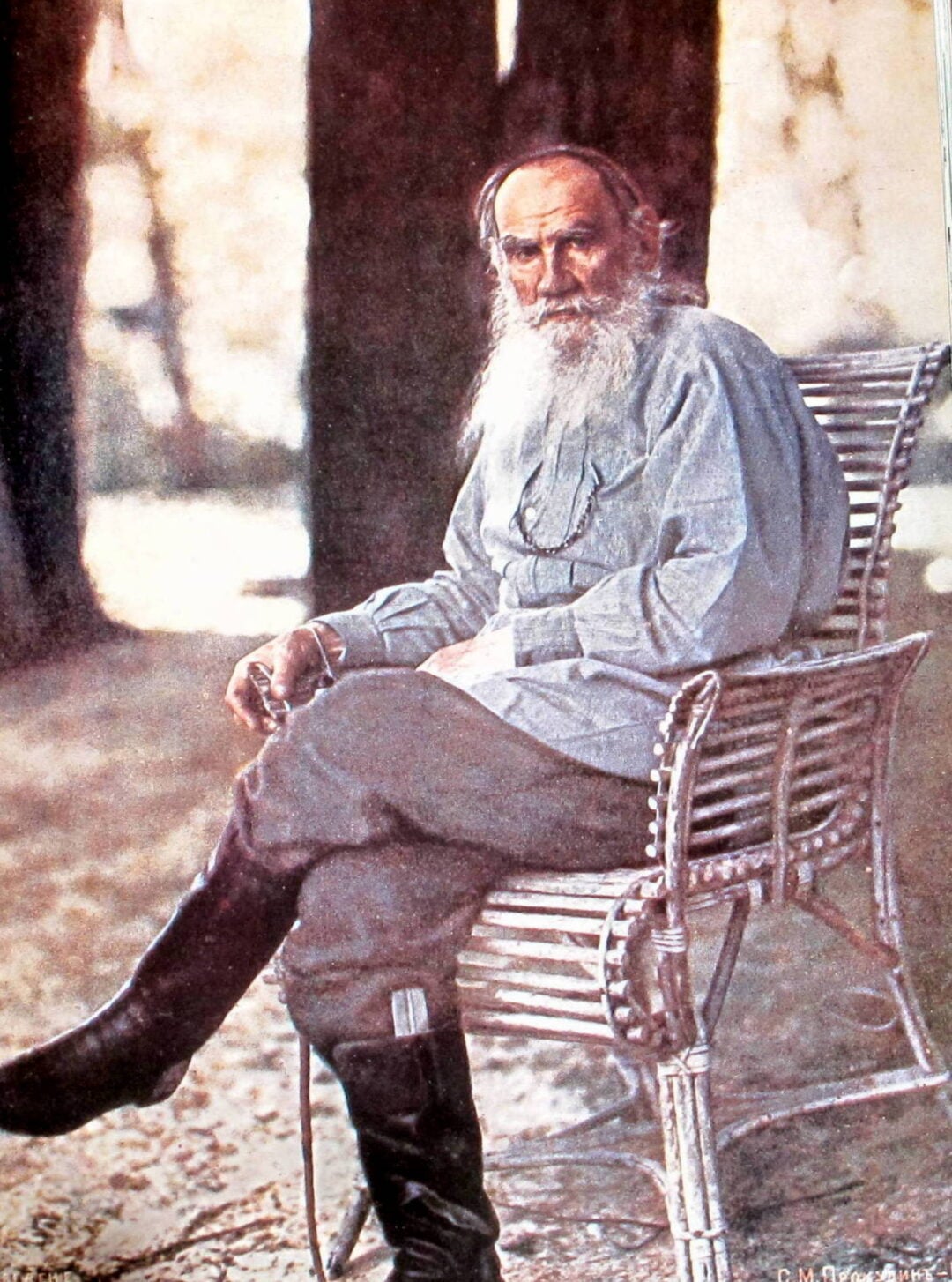 An elderly man with a long white beard sits on a wicker chair outdoors, wearing a light-colored shirt, dark pants, and boots. He gazes calmly at the camera, with trees and soft sunlight in the background.