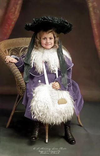 A young girl in a purple coat with white fur trim and a large black hat sits on a wicker chair, smiling. She holds a fluffy white bag and wears lace-up boots. The background is a dark studio backdrop.
