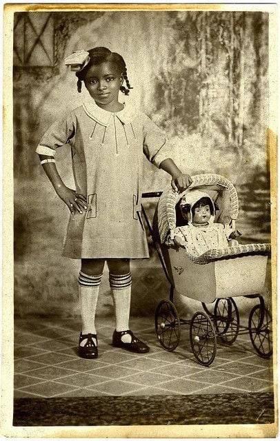 A young Black girl in a dress stands indoors with one hand on her hip and the other on a vintage stroller holding a doll. The photo has a sepia tone and appears to be from the early 20th century.