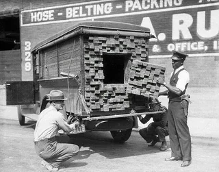 Three men inspect the back of a truck filled with stacked wooden planks concealing an opening, suggesting hidden contraband. The background shows signage for hoses, belting, and packing supplies.