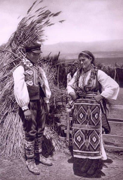 A man and woman in traditional Eastern European folk costumes stand outdoors near tall, bundled wheat stalks, with mountains visible in the background. The woman wears a patterned apron and headscarf; the man wears an embroidered vest and hat.