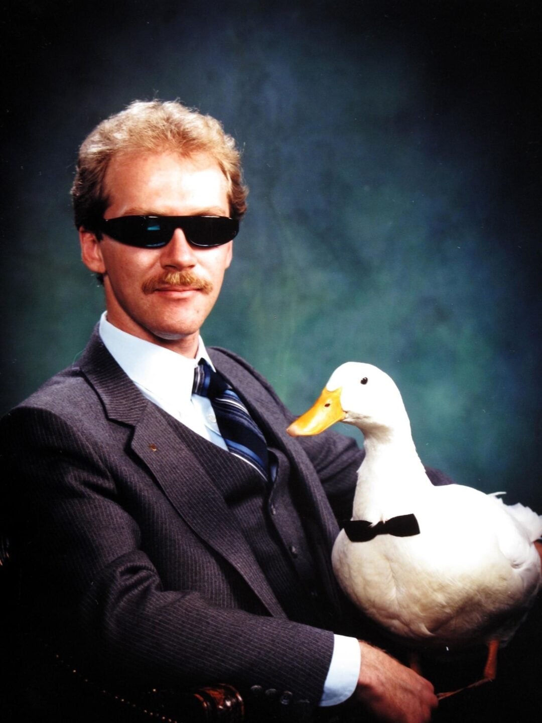 A man in a suit and sunglasses poses for a portrait while holding a white duck wearing a black bow tie, against a dark green studio background.