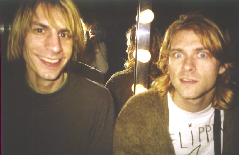 Two young men with long blond hair are sitting in front of a mirror with round lights. One is smiling at the camera, while the other has a surprised expression. Their reflections are visible in the background.