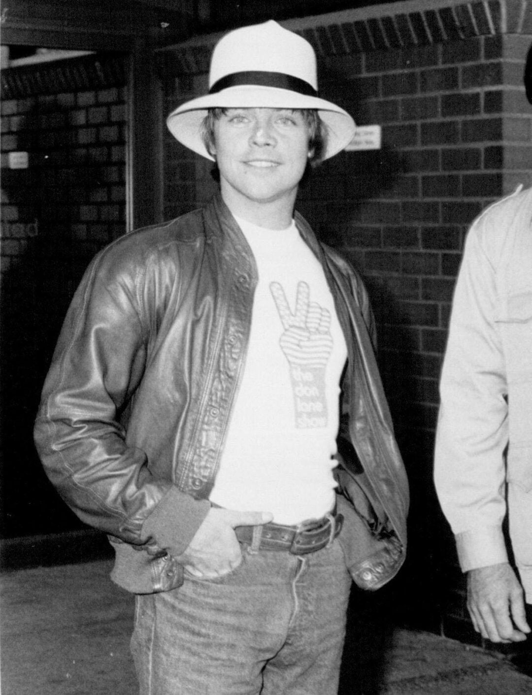 A man wearing a white fedora, leather jacket, and jeans stands smiling. His t-shirt features a large hand making a peace sign. The background includes a brick wall and partial view of another person. The image is in black and white.