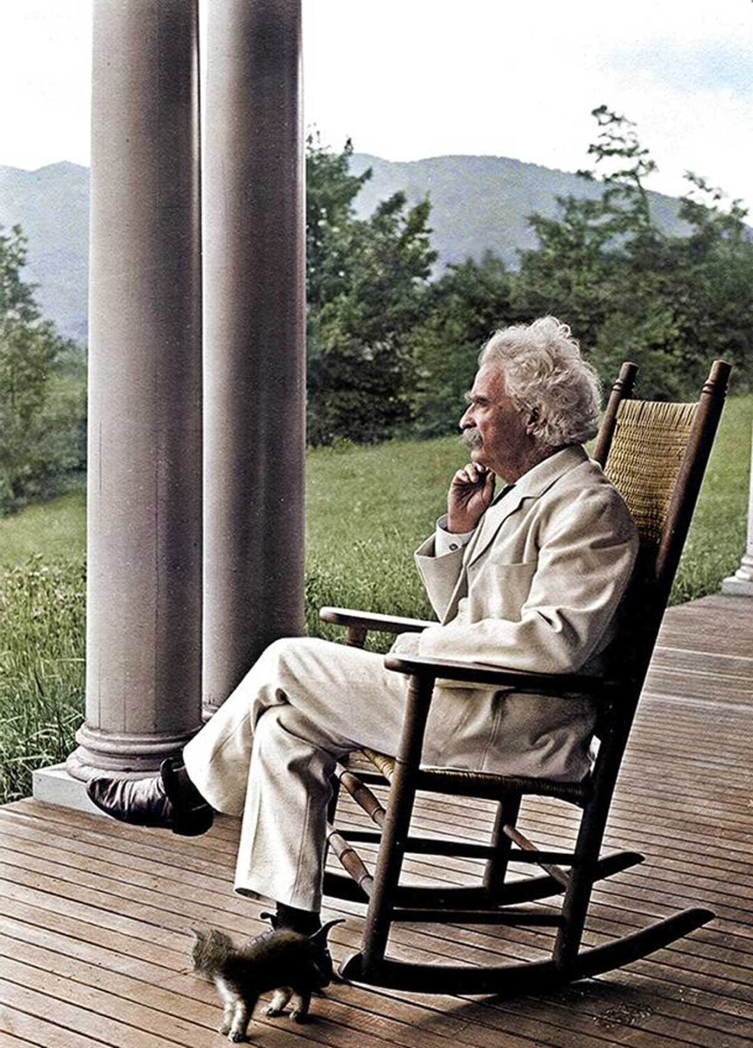 An elderly man with white hair and a white suit sits in a wooden rocking chair on a porch, gazing thoughtfully into the distance. Lush greenery and mountains are visible in the background, with a small cat near his feet.