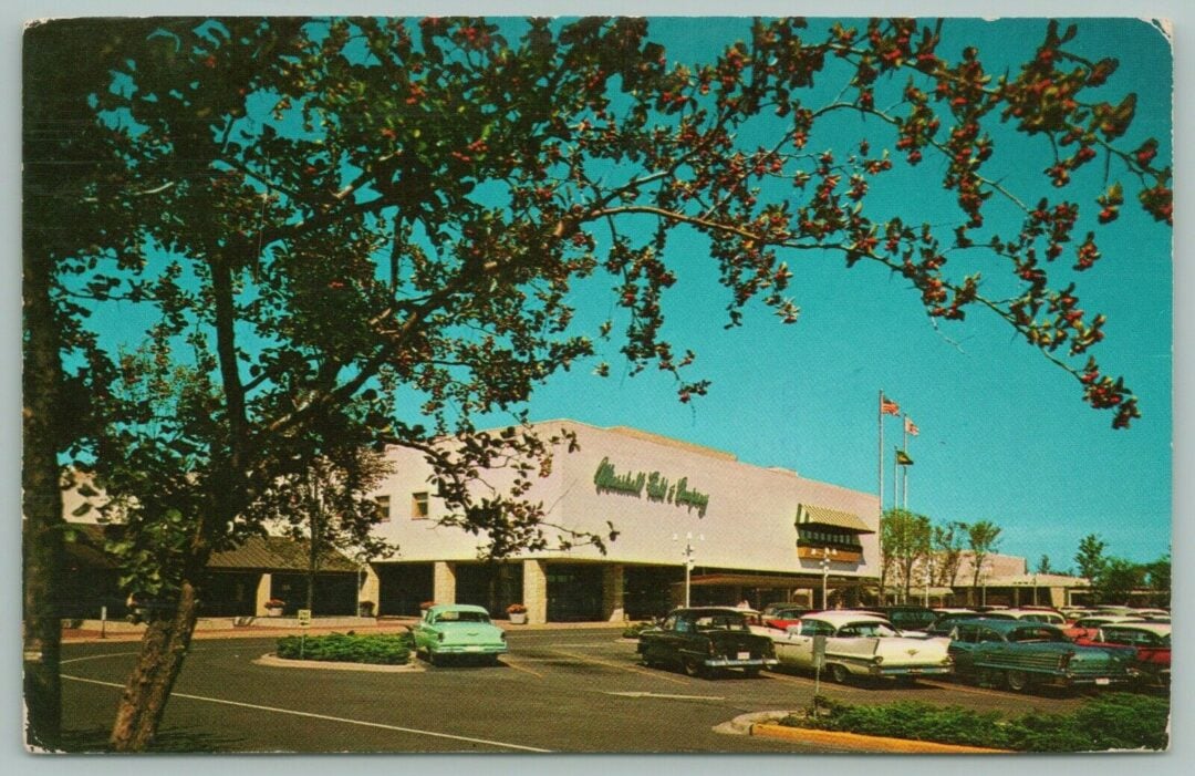A vintage shopping center parking lot with classic cars, a large department store named "Charles V. Loesch," leafy trees in the foreground, and three flagpoles under a bright blue sky.