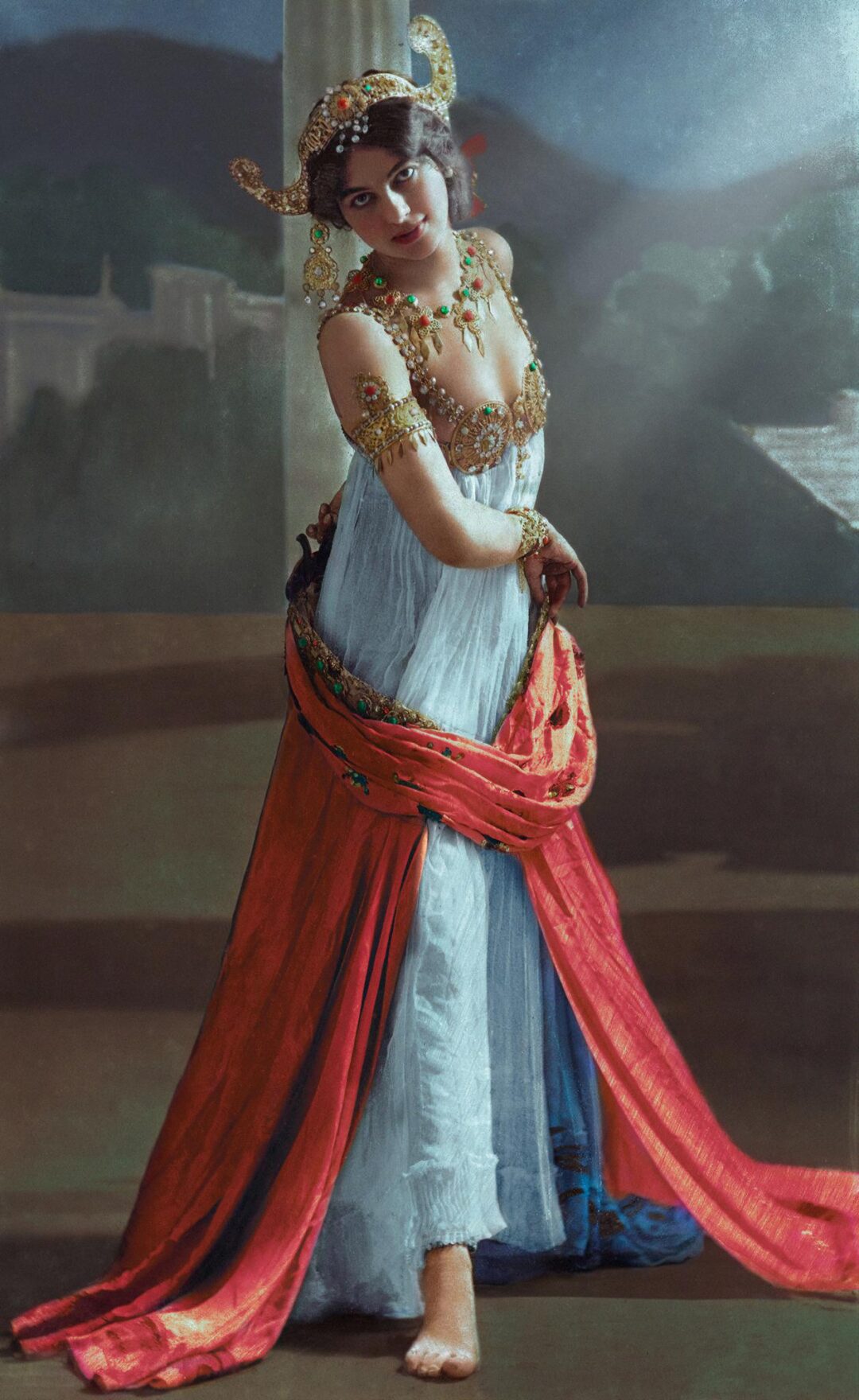A woman in elaborate, beaded costume with gold jewelry, a headdress, and flowing red and blue fabric poses barefoot on a stage with a scenic backdrop of hills and columns.
