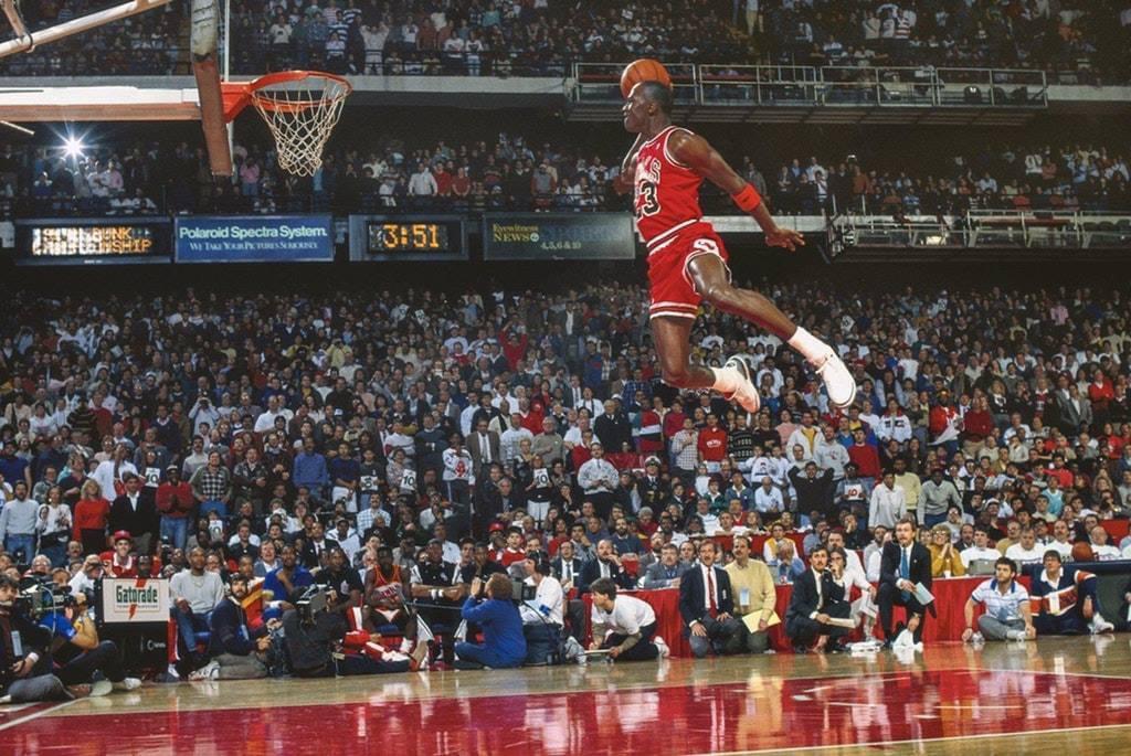 A basketball player in a red uniform leaps toward the hoop for a slam dunk in a packed arena, with fans and photographers watching the dramatic action.