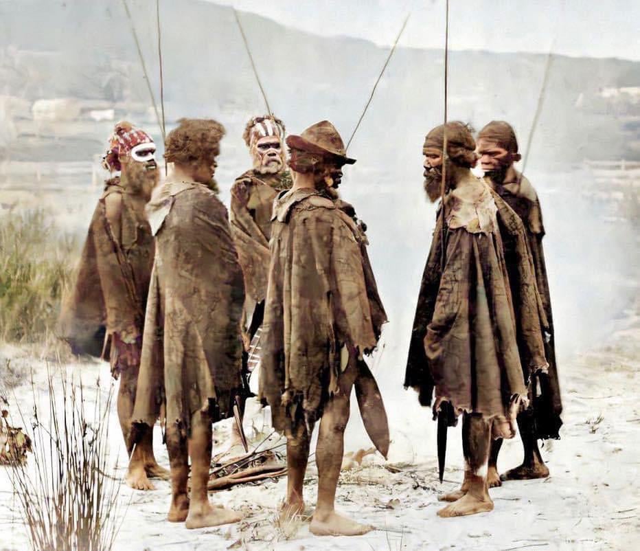Six Indigenous Australian men stand in a circle, wearing traditional cloaks and body paint, with spears in hand, on sandy ground with smoke and natural landscape in the background.