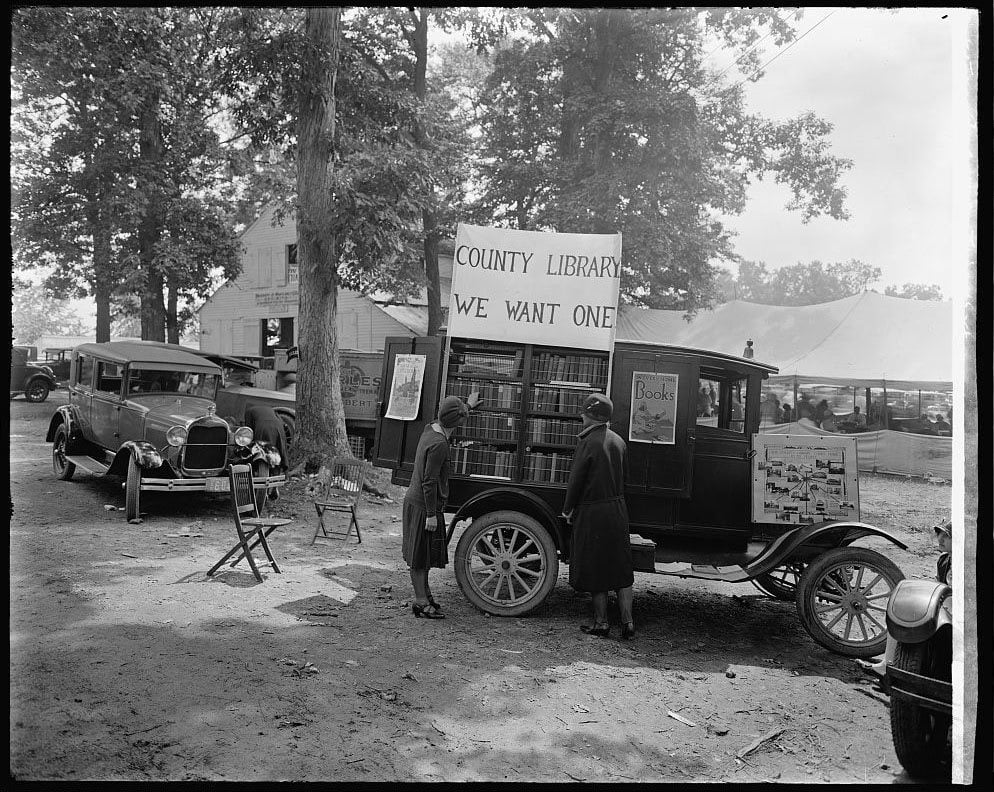 Two women stand by a vintage truck with shelves of books and a sign reading “County Library: We Want One” at an outdoor fair. Old cars and trees are in the background.
