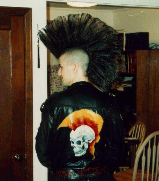A person with a tall, spiked mohawk hairstyle stands with their back to the camera, wearing a black leather jacket featuring a skull and a red mohawk design. They are in a room with wooden furniture and bookshelves.