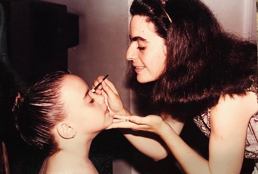A woman applies makeup to a young girl's face with a brush. The girl has her hair pulled back in a bun and her eyes closed, while the woman gently holds her chin and focuses on her work.