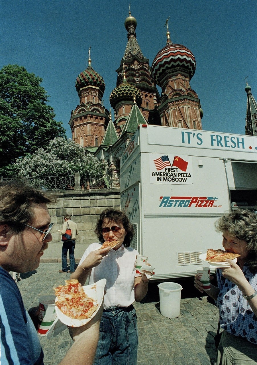 Three people eat pizza in front of a food truck labeled “First American Pizza in Moscow” near Saint Basil’s Cathedral in Moscow, with the iconic colorful domes visible in the background.