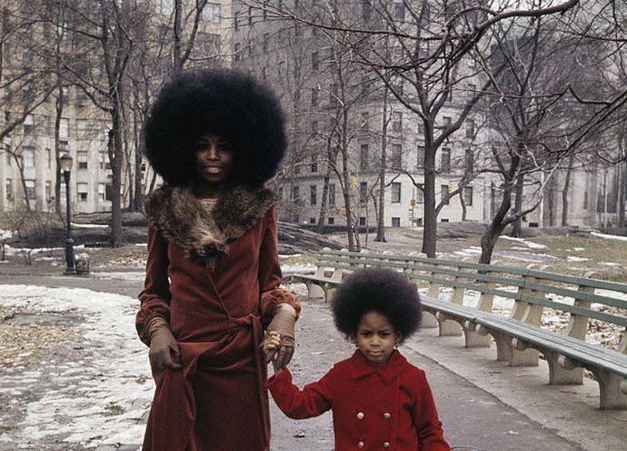 A woman and a young girl, both with large afros and wearing matching red coats, stand hand-in-hand on a park path in winter with bare trees and buildings in the background.