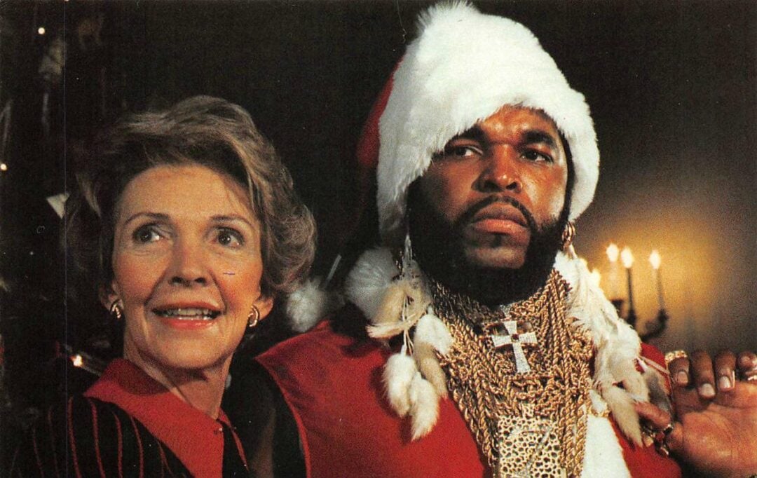 An older woman in a red outfit stands beside a man wearing a Santa hat, red coat, and multiple gold chains, with a dark background and candlelight behind them.
