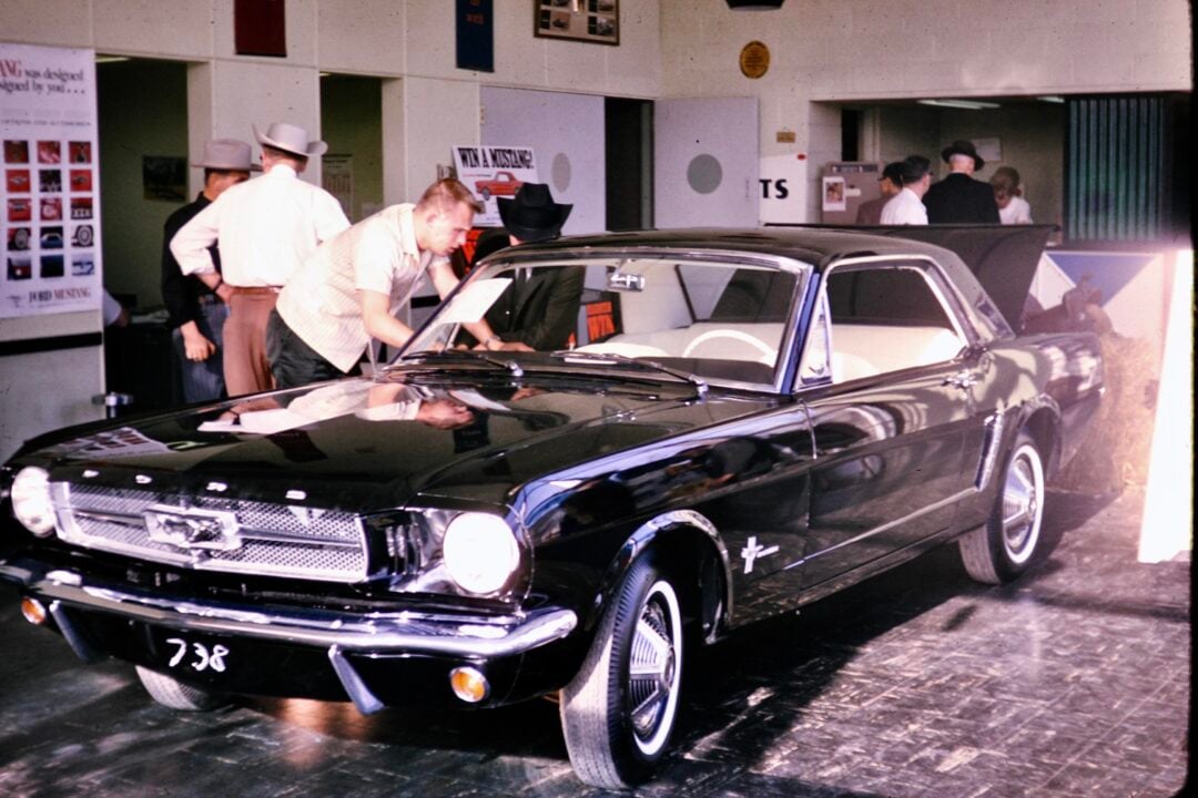 A man inspects a shiny black 1960s Ford Mustang inside a showroom, while several people in hats and suits stand in the background near posters and doors.