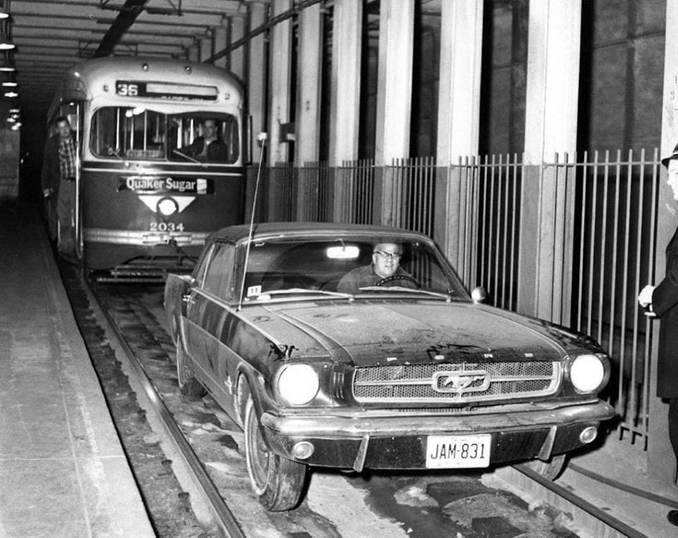A black-and-white photo shows a vintage Ford Mustang stuck on trolley tracks inside a tunnel, with a streetcar labeled "36" stopped closely behind it. A person stands near the Mustang, watching the scene.