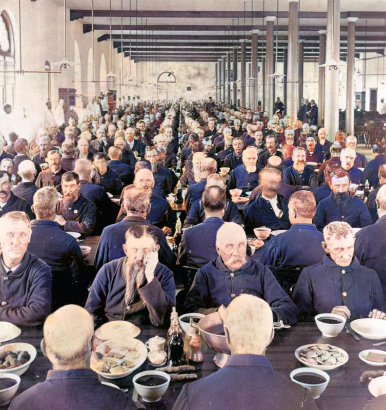 Large hall filled with rows of men in uniform sitting at long tables, eating a meal. The scene is orderly, with food and drinks on the tables, and the background shows more diners and high ceilings with pillars.