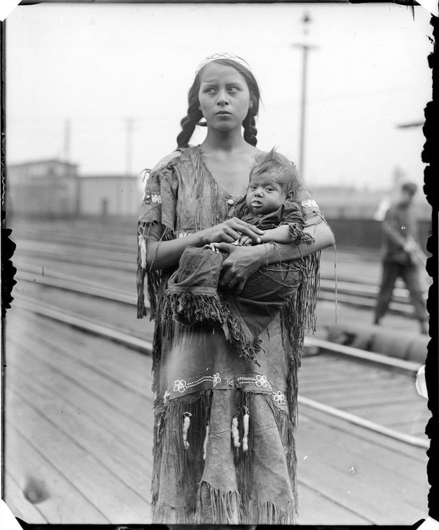 A young Native American woman in traditional dress stands on wooden planks, holding a small child in her arms. The background shows railroad tracks and blurred figures.