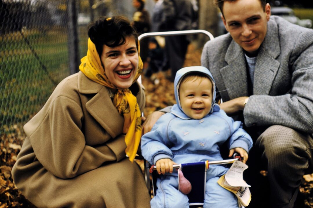 A smiling woman in a tan coat and yellow scarf, a man in a gray suit, and a baby in a blue hooded snowsuit sit outdoors in autumn leaves near a fence. The baby is seated in a stroller, holding a paper.