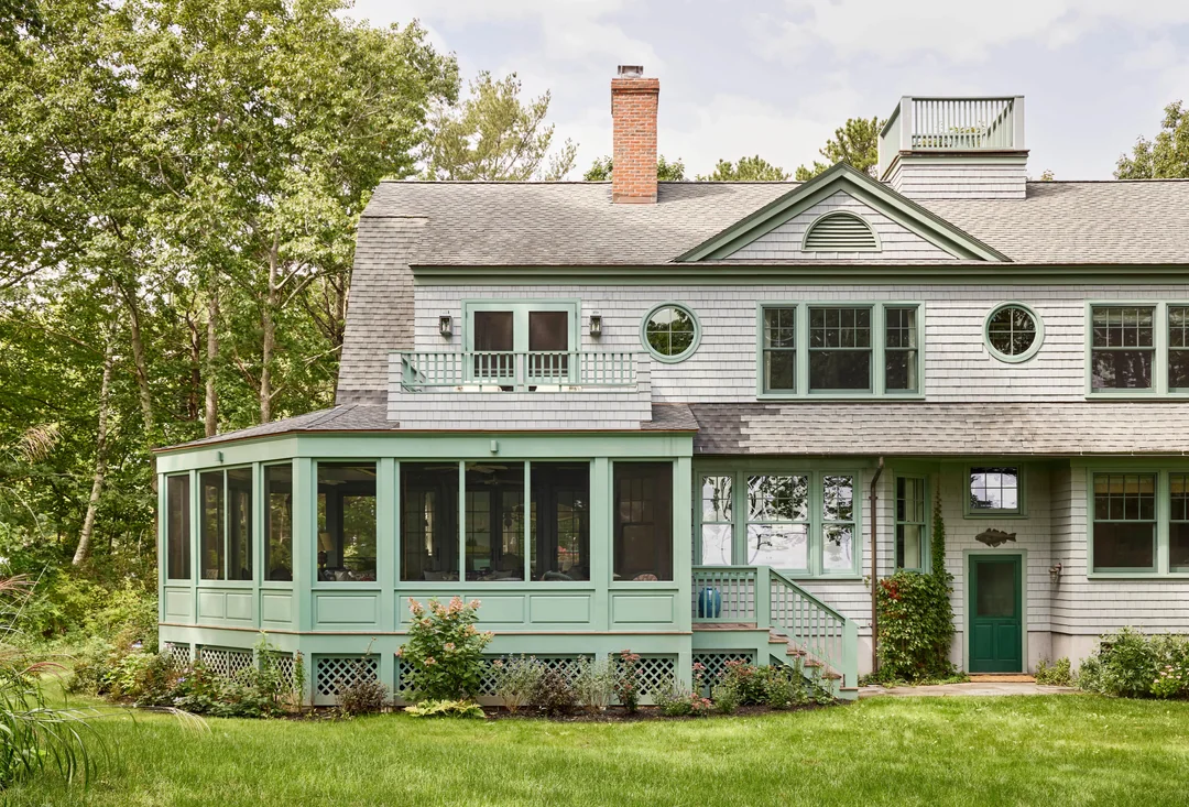 Large, light green house with shingle siding, spacious screened porch, multiple windows, a balcony, green door, chimney, and lush green lawn, surrounded by trees and shrubs.