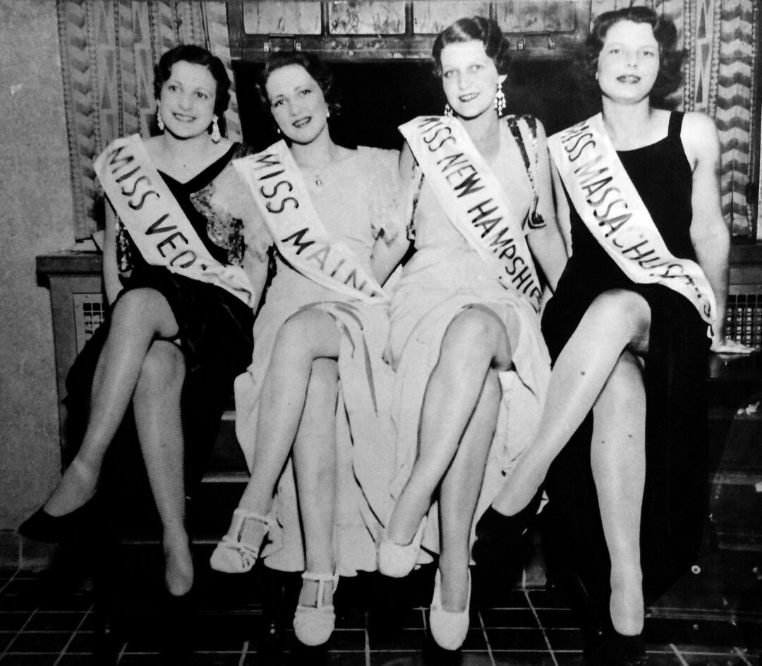 Four women wearing dresses and sashes labeled Miss Vermont, Miss Maine, Miss New Hampshire, and Miss Massachusetts sit on a bench, smiling with their legs crossed in a black and white photo.
