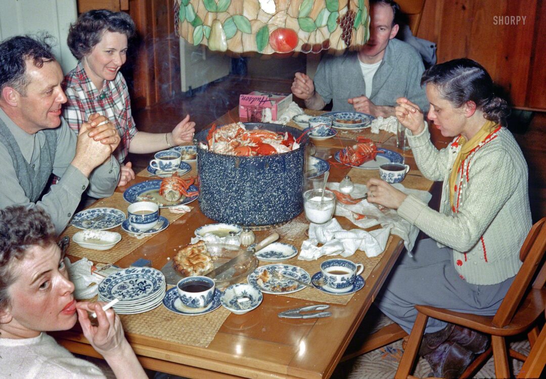 Five adults sit around a dinner table eating crab, with crab shells piled in a large pot. The table is set with patterned dishes, coffee cups, and glasses of milk. The scene appears lively and informal.