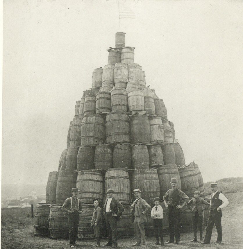 A black-and-white photo of eight people standing in front of a large, pyramid-shaped stack of wooden barrels outdoors. An American flag is on top of the barrels. The setting appears old-fashioned and rural.