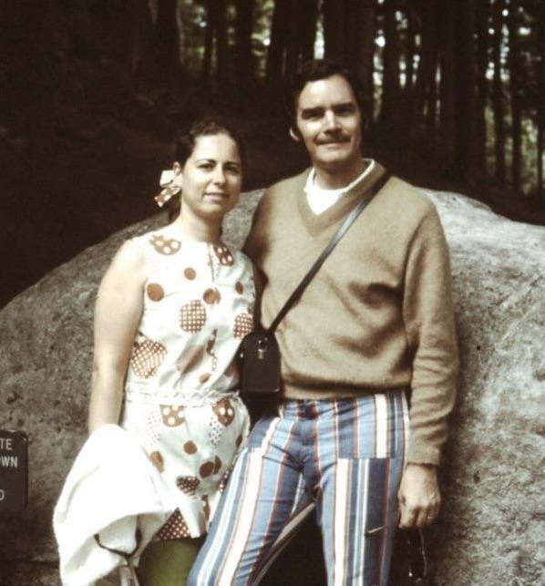 A woman in a patterned dress and a man in a tan sweater with striped pants stand together in front of a large rock, surrounded by trees. They both smile slightly at the camera.