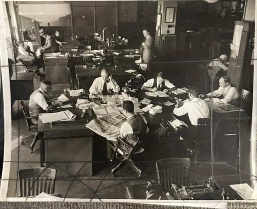 Black-and-white photo of an old-fashioned office with several men in shirts and vests working at desks filled with papers, typewriters, and phones. The room has a busy, collaborative atmosphere.