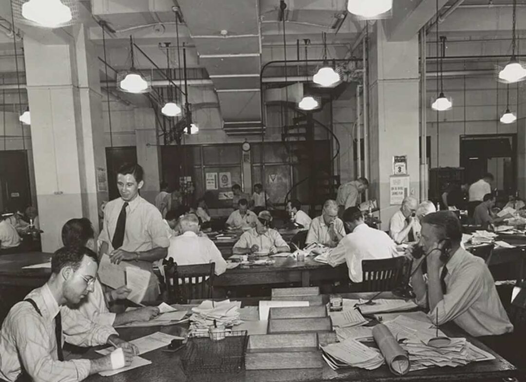 A busy, vintage office scene with men in dress shirts and ties working at large tables covered in papers, typewriters, and stacks of documents under bright overhead lights.
