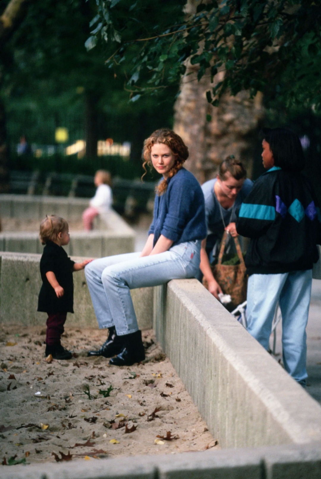 A woman in a blue sweater and light jeans sits on a low concrete wall in a park, looking at the camera. A toddler stands nearby, and two other adults are talking beside her under leafy trees.