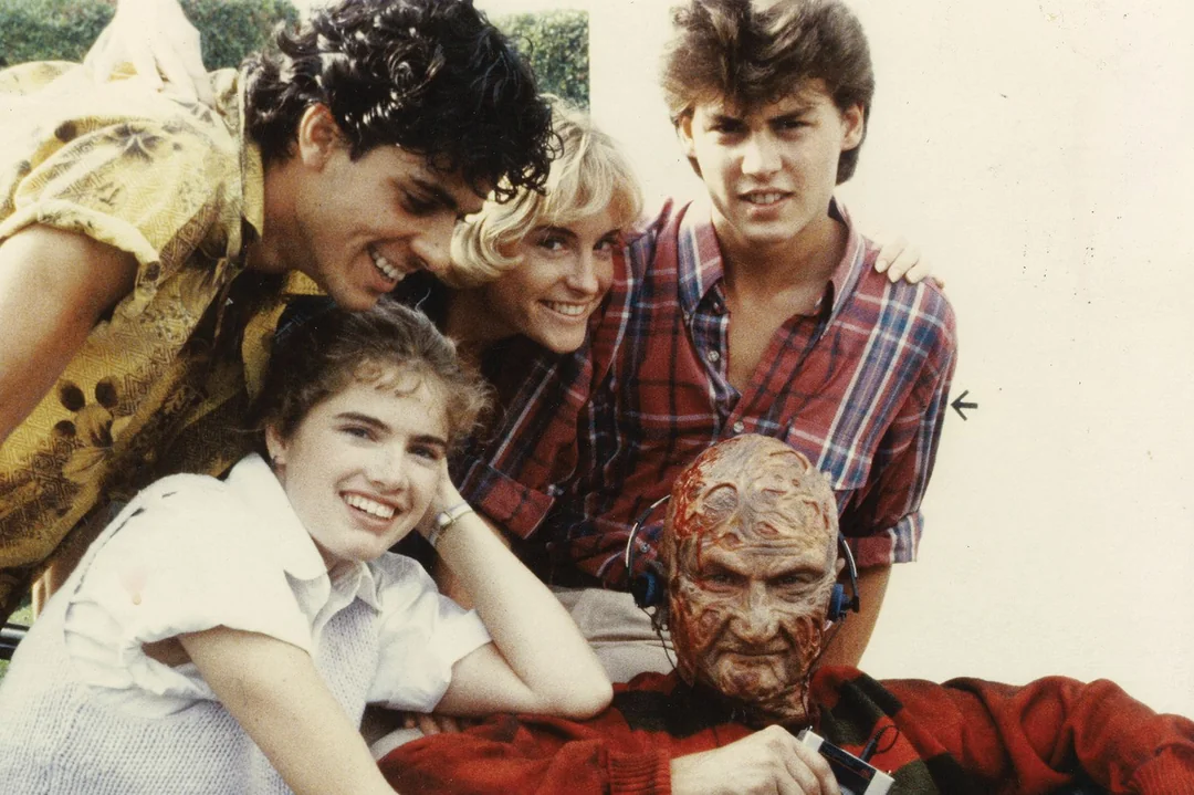 Four smiling young adults pose together behind a person in detailed, gruesome monster makeup with singed skin and wearing headphones. The group is outdoors in daylight, appearing cheerful and friendly.