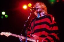A musician with shoulder-length blonde hair plays an electric guitar and sings into a microphone on stage, wearing a red and black striped sweater, under colorful stage lights.