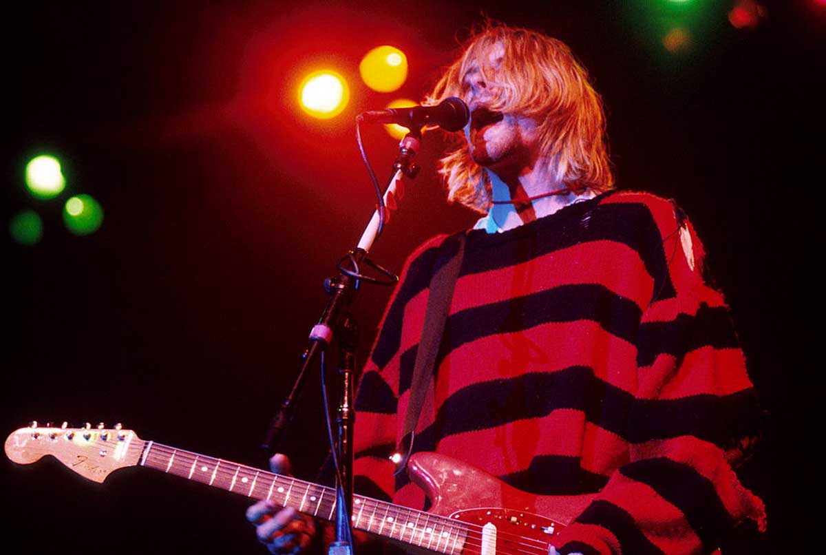 A musician with shoulder-length blonde hair plays an electric guitar and sings into a microphone on stage, wearing a red and black striped sweater, under colorful stage lights.