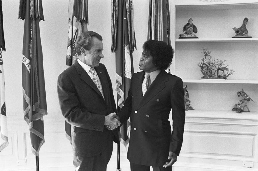 Two men in suits are shaking hands and smiling at each other in front of several flags. They stand in a well-lit room with shelves displaying decorative bird figurines.