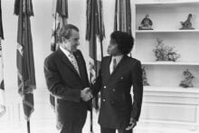 Two men in suits are shaking hands and smiling at each other in front of several flags. They stand in a well-lit room with shelves displaying decorative bird figurines.